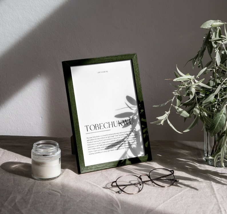 May include: A green framed print with the word "TOBECHUKWU" in black text. The print is on a white background and has a definition of the word below it. The print is sitting on a light brown surface with a white candle and round glasses.