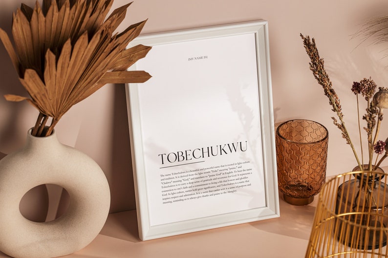 May include: White framed print with the text "MY NAME IS TOBECHUKWU" and a definition of the name. The print is sitting on a table with a vase of dried palm leaves, a brown glass candle holder, and a gold metal vase with dried flowers.