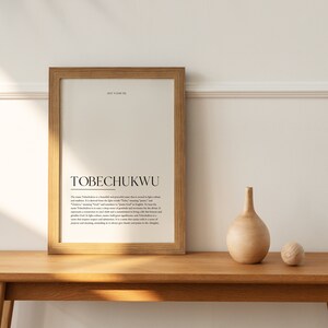 May include: A framed print with the name "TOBECHUKWU" in bold black text. The print explains the meaning of the name, which is a beautiful and powerful name that is meant to inspire calm and wisdom. The name is of Igbo origin and means "God is Great".