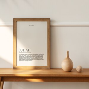 May include: A framed print with the word "JUDAH" in a simple black font. The print has a definition of the name Judah, explaining its meaning and significance. The print is on a wooden table with a wooden vase and a wooden ball.