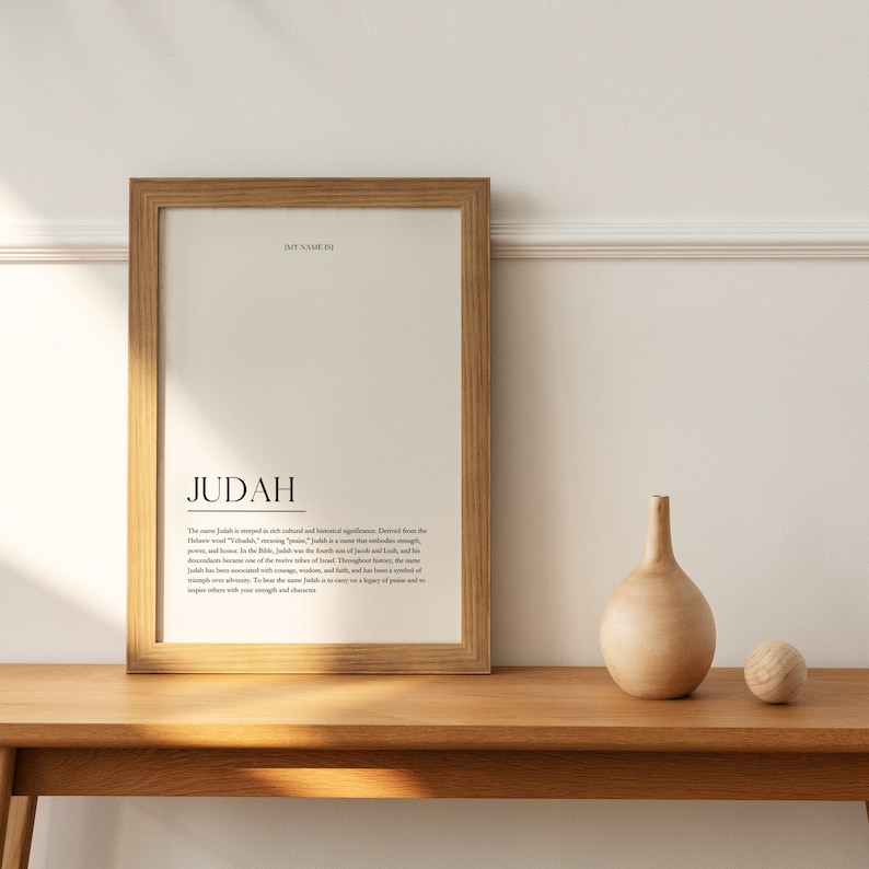 May include: A framed print with the word "JUDAH" in a simple black font. The print has a definition of the name Judah, explaining its meaning and significance. The print is on a wooden table with a wooden vase and a wooden ball.