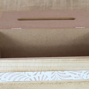 May include: A rectangular wooden tissue box cover with a hinged lid featuring a central slot. The exterior displays a light natural wood grain finish accented by a horizontal white band decorated with a repeating tan leaf pattern across the front.