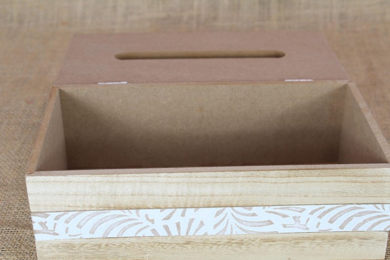 May include: A rectangular wooden tissue box cover with a hinged lid featuring a central slot. The exterior displays a light natural wood grain finish accented by a horizontal white band decorated with a repeating tan leaf pattern across the front.