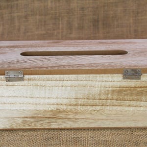 May include: A rectangular wooden tissue box cover with a light brown natural wood grain finish. The top features a centered elongated oval opening for dispensing tissues, and the lid is attached to the base with two silver metal hinges.