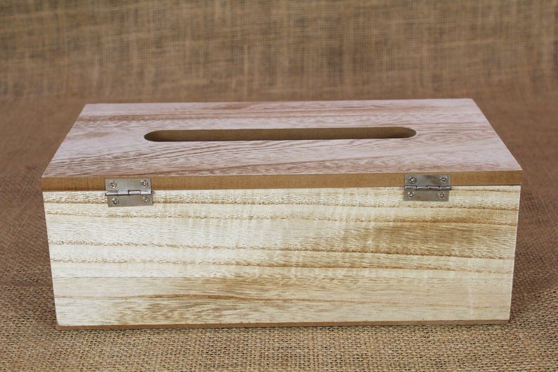May include: A rectangular wooden tissue box cover with a light brown natural wood grain finish. The top features a centered elongated oval opening for dispensing tissues, and the lid is attached to the base with two silver metal hinges.