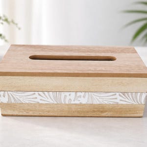 May include: A rectangular wooden tissue box cover featuring a natural light brown wood grain finish on the top and sides, with a horizontal band across the center displaying a white and light beige leaf pattern design and a top slot opening.
