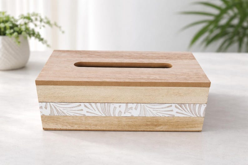 May include: A rectangular wooden tissue box cover featuring a natural light brown wood grain finish on the top and sides, with a horizontal band across the center displaying a white and light beige leaf pattern design and a top slot opening.