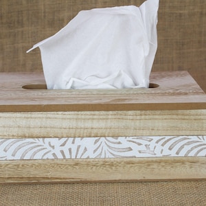 May include: A rectangular wooden tissue box cover with a light brown natural wood grain finish. The front features a horizontal white band decorated with a repeating leaf pattern. A single white tissue is pulled through the top opening.