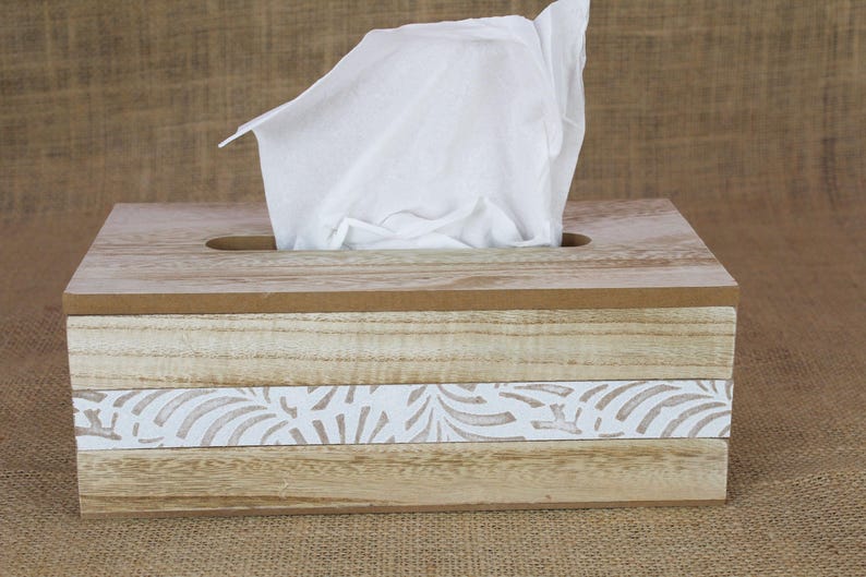 May include: A rectangular wooden tissue box cover with a light brown natural wood grain finish. The front features a horizontal white band decorated with a repeating leaf pattern. A single white tissue is pulled through the top opening.