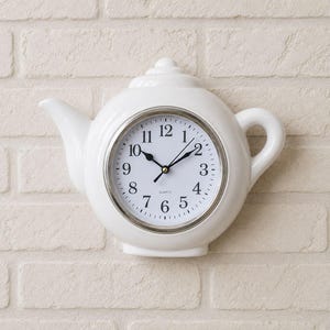 May include: A white ceramic wall clock shaped like a teapot. The clock face is white with black Arabic numerals, black hour and minute hands, and a thin silver-toned metal bezel. The word QUARTZ is printed in small black letters at the bottom.