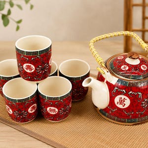 May include: A ceramic tea set featuring a teapot with a woven bamboo handle and five matching cups. The pieces are decorated in a vibrant red with intricate black floral patterns, a geometric border, and white circular medallions containing Chinese characters.