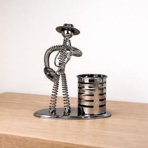 Op de afbeelding: Een zilverkleurig metalen beeldje van een muzikant met een hoed die een hoorn bespeelt, opgebouwd uit spiraalveren en metalen onderdelen. Het figuur staat op een ovale voet naast een cilindervormige pennenhouder met horizontale sleuven.