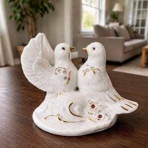Puede incluir: Una figura de cerámica blanca de dos palomas posadas juntas sobre una base. Las aves presentan detalles pintados en oro en las alas y picos, con pedrería transparente incrustada en sus pechos y dos pequeñas piedras rojas en la base cerca de las colas.