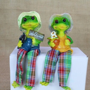 Könnte beinhalten: Zwei grüne Froschfiguren mit Hüten und karierten Hosen sitzen auf einer weißen Oberfläche. Ein Frosch hält ein Schild mit der Aufschrift "Willkommen".