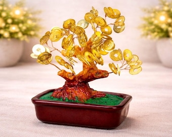 May include: A decorative bonsai tree with a brown trunk and branches adorned with gold-coloured coin-shaped ornaments. The tree sits in a rectangular, maroon-coloured pot filled with green moss. The background features blurred white vases.