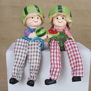 May include: Two ceramic figurines of people wearing watermelon hats and holding watermelon slices. The figurines are sitting on a white shelf with red and white checkered pants.