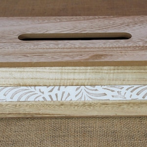May include: A rectangular wooden tissue box cover featuring a light brown wood grain top with a central slot. The sides consist of horizontal light wood planks with a central white band decorated with a repeating light brown leaf pattern.