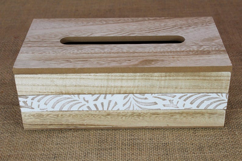 May include: A rectangular wooden tissue box cover featuring a light brown wood grain top with a central slot. The sides consist of horizontal light wood planks with a central white band decorated with a repeating light brown leaf pattern.