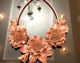 Peut inclure: Un collier en cuir rose gold avec trois grandes fleurs en cuir sculptées. Les fleurs ont un design texturé détaillé et sont disposées en grappe.