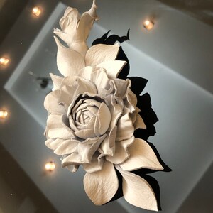 May include: A white leather rose with leaves, crafted in a realistic style. The rose is a single bloom with multiple petals and a center bud. The leaves are detailed and textured.