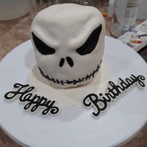 May include: A white cake decorated to resemble Jack Skellington's face from The Nightmare Before Christmas. The cake has black eyes, a stitched mouth, and the words "Happy Birthday" in black script on a white plate.