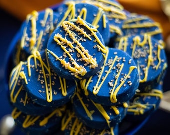 May include: A close-up shot of a pile of blue-frosted cookies in a blue bowl. Each cookie is decorated with yellow drizzle and gold sprinkles. The cookies are arranged in a visually appealing way, suggesting a dessert or treat.