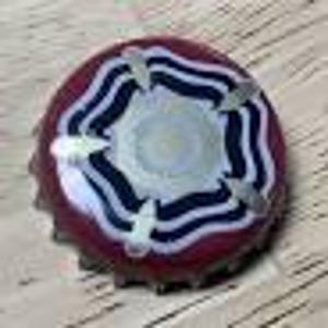 May include: A decorative bottle cap with a burgundy base and a white and navy blue floral design. The cap's design features a central circle surrounded by wavy lines and silver accents. The cap is approximately 2.5 cm in diameter.