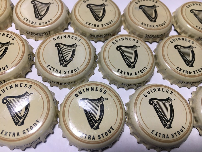 May include: A collection of Guinness Extra Stout bottle caps. Each cap features the Guinness logo, a black harp, and the text "EXTRA STOUT". The caps are cream-colored with a gold border and black text.