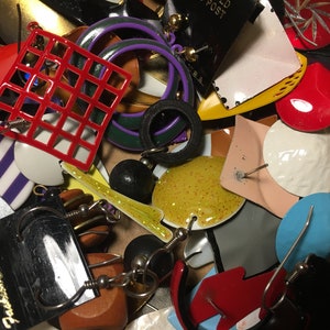 May include: A collection of colorful and unique earrings, including a red grid pattern, a yellow glitter circle, a white circle with a textured surface, and a blue circle. The earrings are displayed on a brown surface with a black tag that reads "Mostly Fashion Earrings".