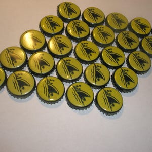 May include: A collection of yellow bottle caps with a black design featuring a stylized ship and waves. The caps are arranged in a diamond shape, with a black rim and a glossy finish. The caps are likely for beer or soda bottles.