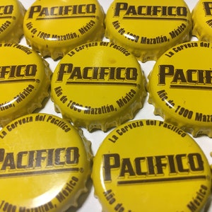 May include: A collection of yellow Pacifico beer bottle caps. Each cap features the Pacifico logo in brown, along with text in Spanish. The caps are from Mazatlán, Mexico, and the year 1900 is also visible.