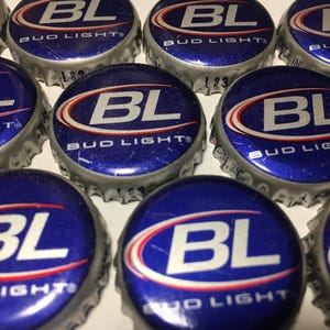 May include: Close-up of several blue and silver Bud Light bottle caps. Each cap features the letters "BL" in white, with a red and white curved design, and the words "Bud Light" in white. The caps are arranged in a close, overlapping pattern.