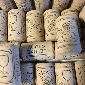 May include: Close-up of a pile of cork wine bottle stoppers with the text "AGGLO DELUXE" printed on them. The stoppers feature a design of a wine glass and grapes.