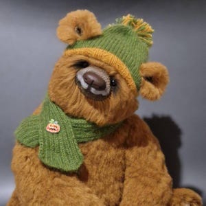 May include: A brown plush teddy bear with a dark brown nose and mouth area. The bear wears a green and yellow knit beanie with a pom-pom on top and a matching green knit scarf. A small orange pumpkin-shaped pin is attached to the scarf.