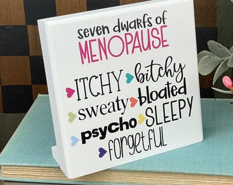 May include: A white wooden block with the text "seven dwarfs of MENOPAUSE" in pink and black. The words "ITCHY, bitchy, sweaty, bloated, psycho, sleepy, forgetful" are listed below in various colors.