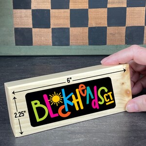 Pu&ograve; includere: Un blocco di legno rettangolare, lungo 15,2 cm e largo 5,7 cm, con la scritta "BLCKHEADS CT" in lettere colorate su sfondo nero. Il blocco &egrave; tenuto in mano e lo sfondo include un motivo a scacchiera.