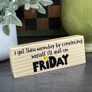 May include: A rectangular wooden block with the text "I get thru monday by convincing myself I'll quit on FRIDAY" in black letters. A yellow smiley face with a wink is between "myself" and "FRIDAY". The block is made of light-colored wood.