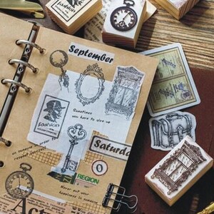 Op de afbeelding: Een bruin lederen gebonden dagboek met metalen ringen en verschillende decoratieve stempels en stickers. De stempels en stickers bevatten afbeeldingen van een sleutel, een raam, een klok, een koffer en een man met een hoge hoed. Het dagboek heeft het woord "September" op de omslag.