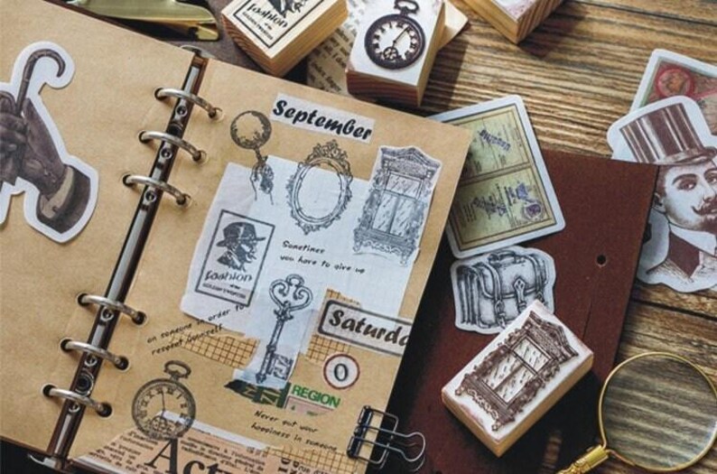 Op de afbeelding: Een bruin lederen gebonden dagboek met metalen ringen en verschillende decoratieve stempels en stickers. De stempels en stickers bevatten afbeeldingen van een sleutel, een raam, een klok, een koffer en een man met een hoge hoed. Het dagboek heeft het woord "September" op de omslag.