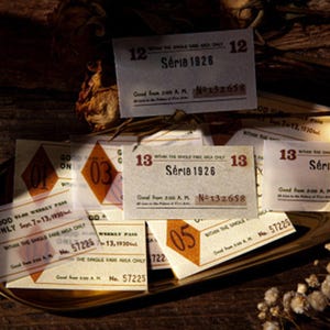 Può includere: Una collezione di biglietti in stile vintage con la scritta "Série 1926" e numeri. I biglietti sono disposti su un vassoio dorato, alcuni con dettagli a forma di diamante arancione e testo aggiuntivo.