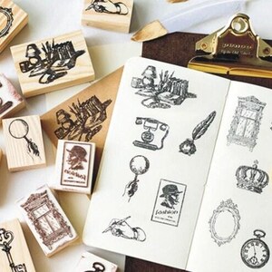 Op de afbeelding: Een verzameling houten stempels met verschillende vintage ontwerpen, waaronder een kroon, een vergrootglas, een veer, een sleutel, een raam, een telefoon en een boek. De stempels zijn gerangschikt op een wit oppervlak met een notitieboekje en een vergrootglas.