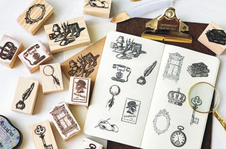 Op de afbeelding: Een verzameling houten stempels met verschillende vintage ontwerpen, waaronder een kroon, een vergrootglas, een veer, een sleutel, een raam, een telefoon en een boek. De stempels zijn gerangschikt op een wit oppervlak met een notitieboekje en een vergrootglas.