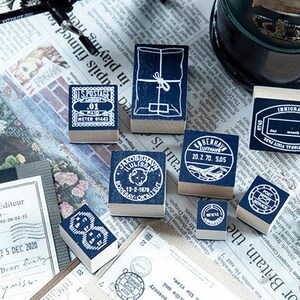 May include: A collection of vintage-style rubber stamps with blue ink pads. The stamps feature various designs, including postage details, package illustrations, and travel-related imagery. They are arranged on a newspaper background.