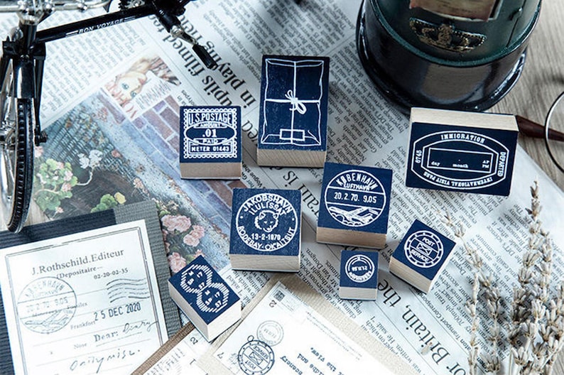 May include: A collection of vintage-style rubber stamps with blue ink pads. The stamps feature various designs, including postage details, package illustrations, and travel-related imagery. They are arranged on a newspaper background.