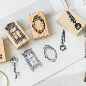 Op de afbeelding: Set van zes houten stempels met vintage ontwerpen. De stempels tonen een sleutel, een raam, een spiegel, een veer en een inktpot, en een tweede raam. De stempels liggen op een wit oppervlak met een gouden veer.