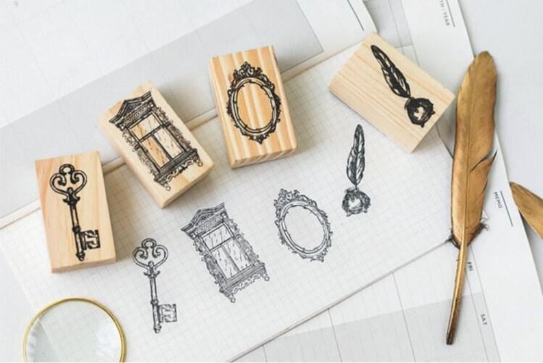 Op de afbeelding: Set van zes houten stempels met vintage ontwerpen. De stempels tonen een sleutel, een raam, een spiegel, een veer en een inktpot, en een tweede raam. De stempels liggen op een wit oppervlak met een gouden veer.