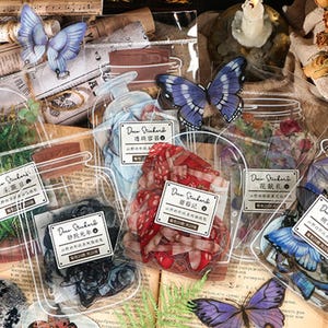 May include: An assortment of decorative stickers in clear plastic containers. The stickers feature butterflies, greenery, and other botanical elements. Each container has a small label with text. The overall aesthetic is vintage and whimsical.