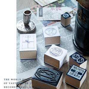 May include: A collection of vintage postage stamps and a silver ink stamp holder are arranged on a newspaper. The stamps feature various designs, including postal markings and text. The wooden bases and detailed designs create a nostalgic aesthetic.