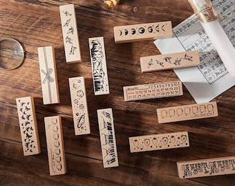 May include: A collection of wooden stamps with various designs, including a calendar, a moon, a forest, a bow, planets, strawberries, and a floral pattern. The stamps are on a wooden surface.