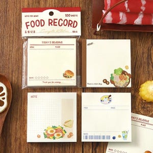 May include: A set of food-themed memo pads with various designs. The pads feature illustrations of burgers, sandwiches, and other dishes. The top pad reads "FOOD RECORD" and "100 SHEETS".